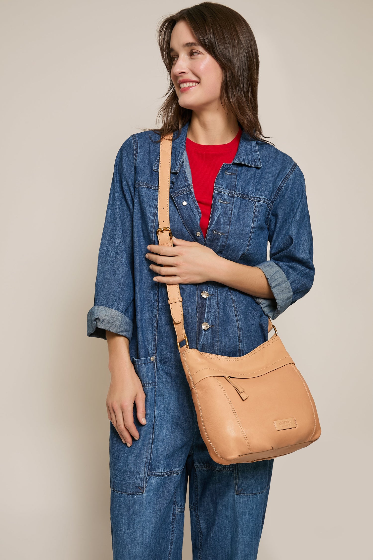 A person in a denim jumpsuit and red top smiles while holding a tan leather crossbody bag. The background is plain and neutral.