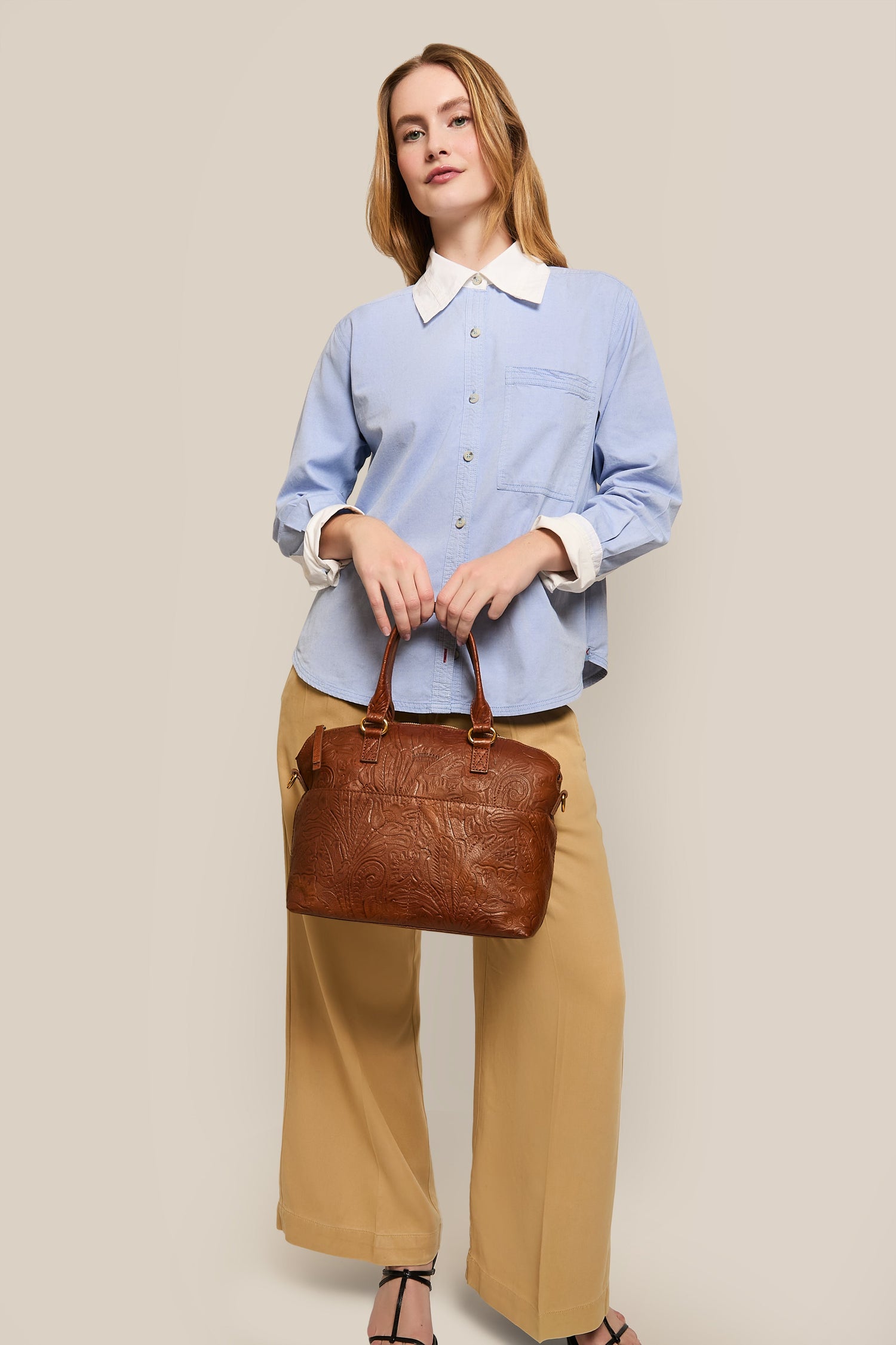 A person in a light blue shirt and beige wide-leg pants stands against a neutral background, holding a brown leather handbag.