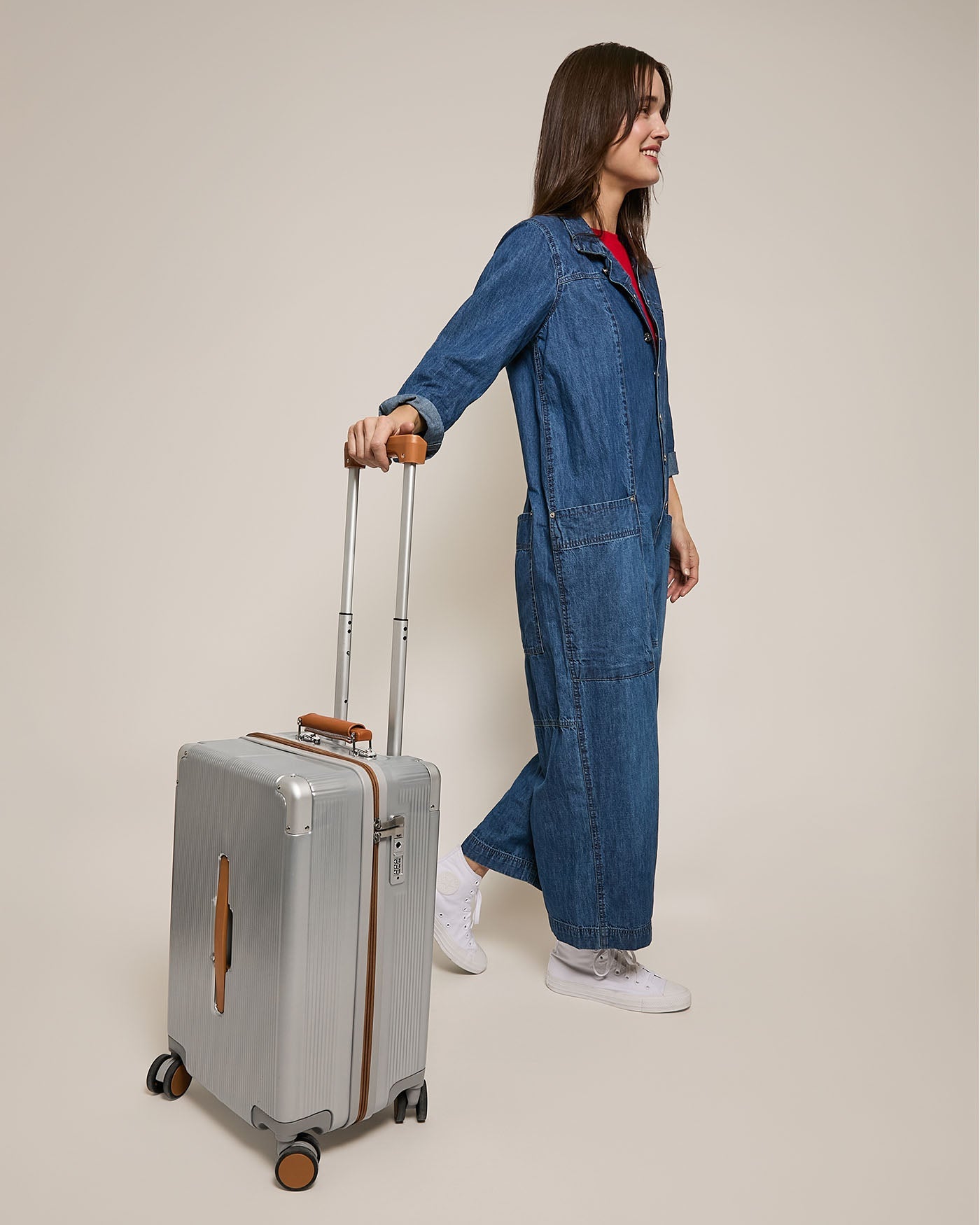 Person in a blue denim jumpsuit and white sneakers pulling a green suitcase with brown handles. The background is plain beige.