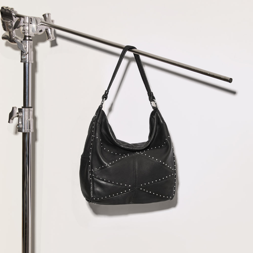 The American Leather Co Locke Ziptop Studded hobo in black hanging on a photography stand on a white wall.