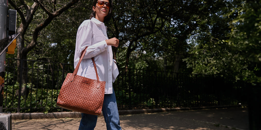 A woman walking by a park while carrying the American Leather Co Sandy Woven Satchel in brandy.