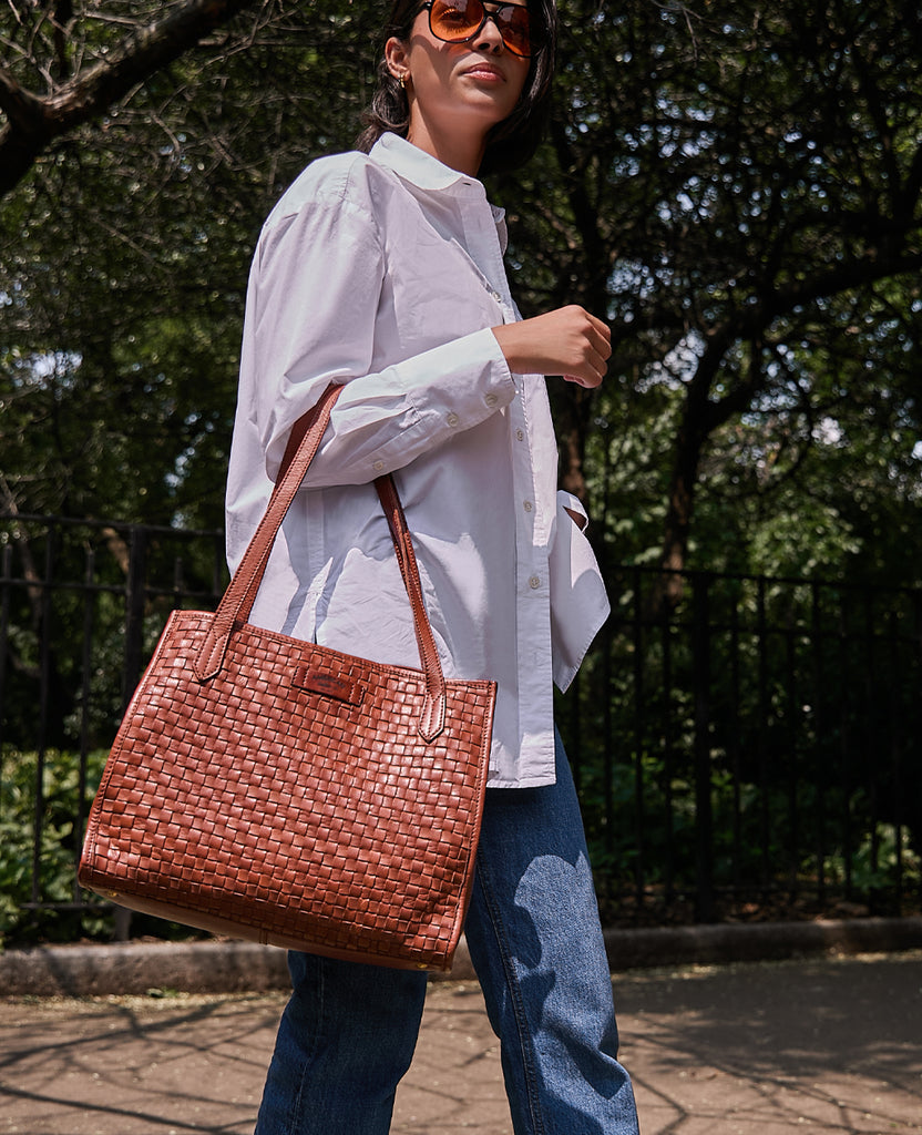 A woman walking by a park while carrying the American Leather Co Sandy Woven Satchel in brandy.