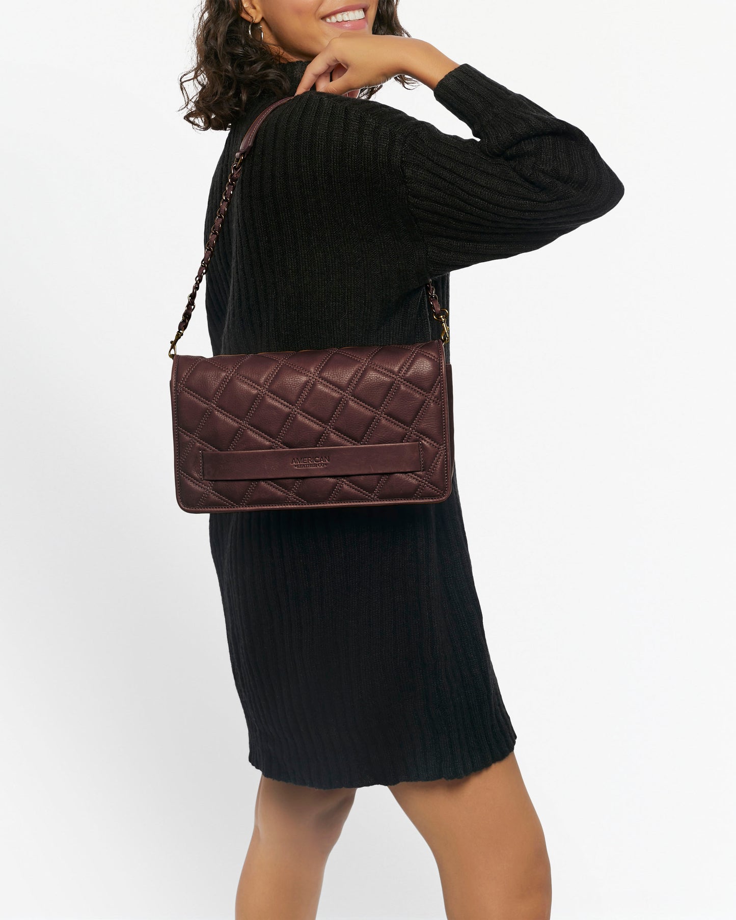 A person in a black ribbed sweater dress poses with an Emerson Quilted Convertible Shoulder by American Leather Co., showcasing its chain strap. The maroon quilted leather pops as they angle to the side against a plain white backdrop.