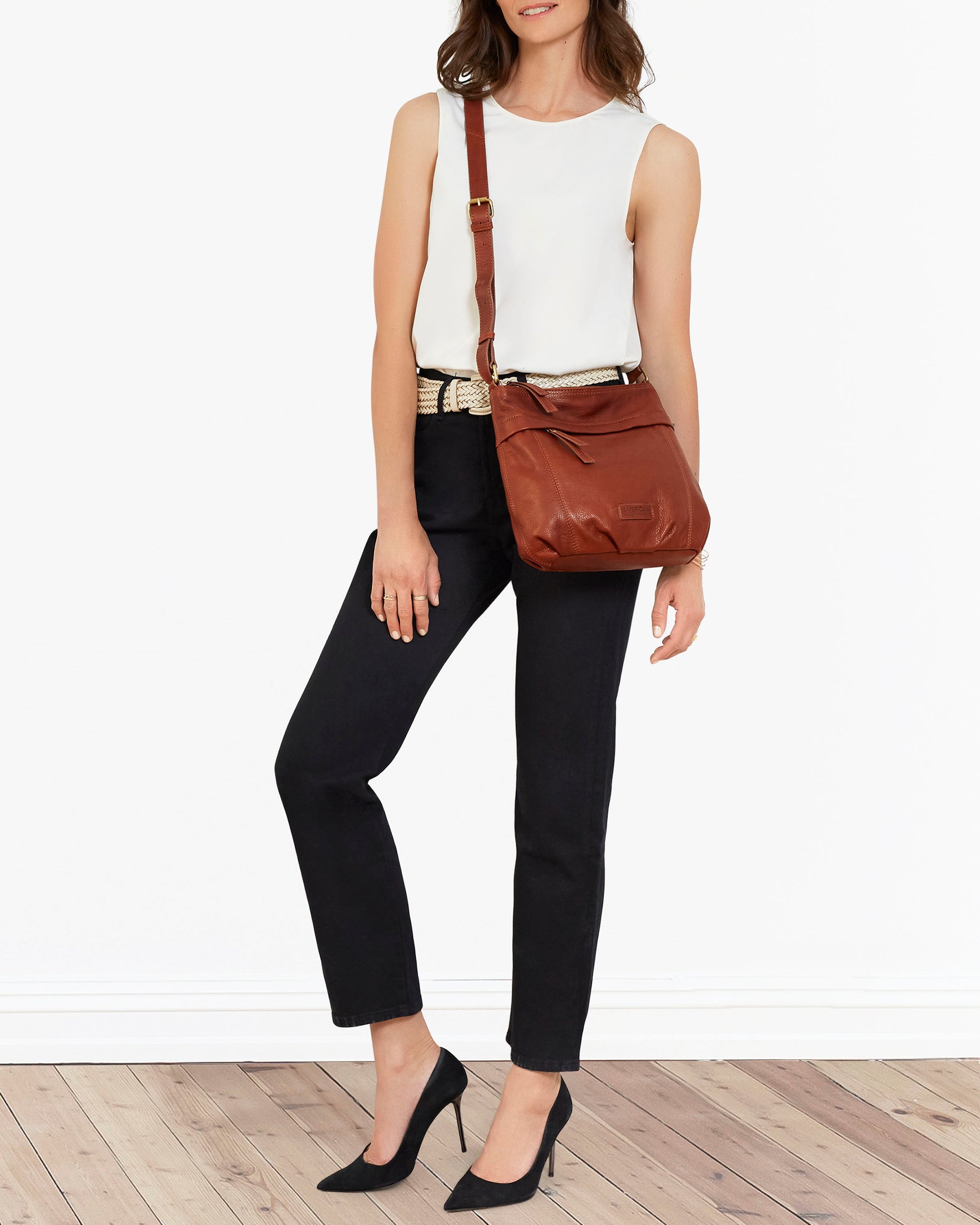 A woman stands against a white background wearing a sleeveless white top, black pants, and black heels. She carries the Acorn Double Entry Crossbody by American Leather Co., crafted from genuine glove leather with antiqued hardware, and has a light-colored belt. The floor is wooden.