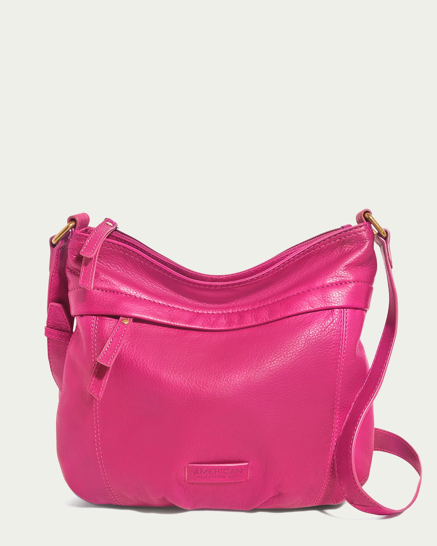 Introducing the Acorn Double Entry Crossbody by American Leather Co., a vibrant pink bag meticulously crafted from genuine glove leather with a textured finish. It boasts a zippered top, an adjustable strap adorned with antiqued brass hardware, and features a small logo patch sewn onto the front.