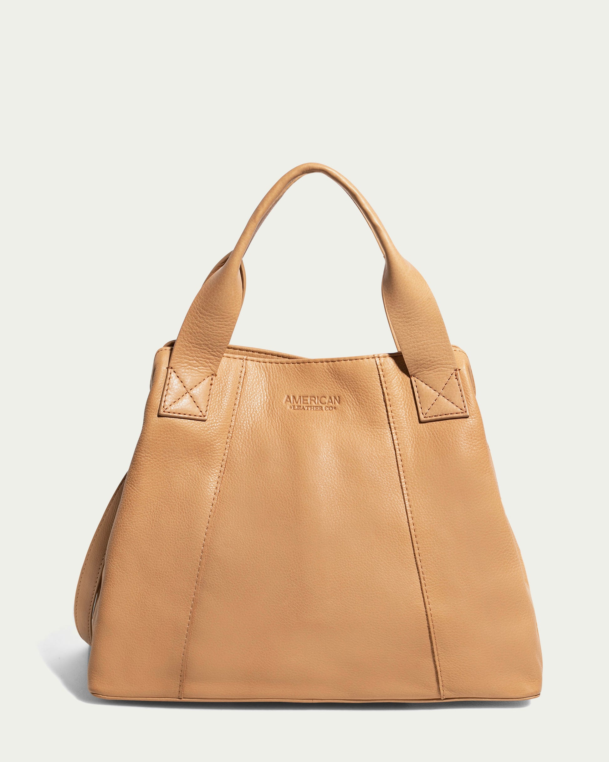 The Ada Triple Entry Satchel by American Leather Co. is a tan leather handbag made from genuine American leather. It features two sturdy handles, a subtle embossed logo on the front, and a plain cream background. The structured satchel has a rectangular shape and visible stitching for added detail.