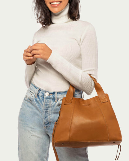 A person wearing a light gray turtleneck and light-wash jeans is smiling and holding the Ada Triple Entry Satchel from American Leather Co. with both hands. The structured bag, made from Genuine American Leather, features two short handles and an adjustable crossbody strap, complementing the person's long, dark hair visible against the plain, light-colored background.