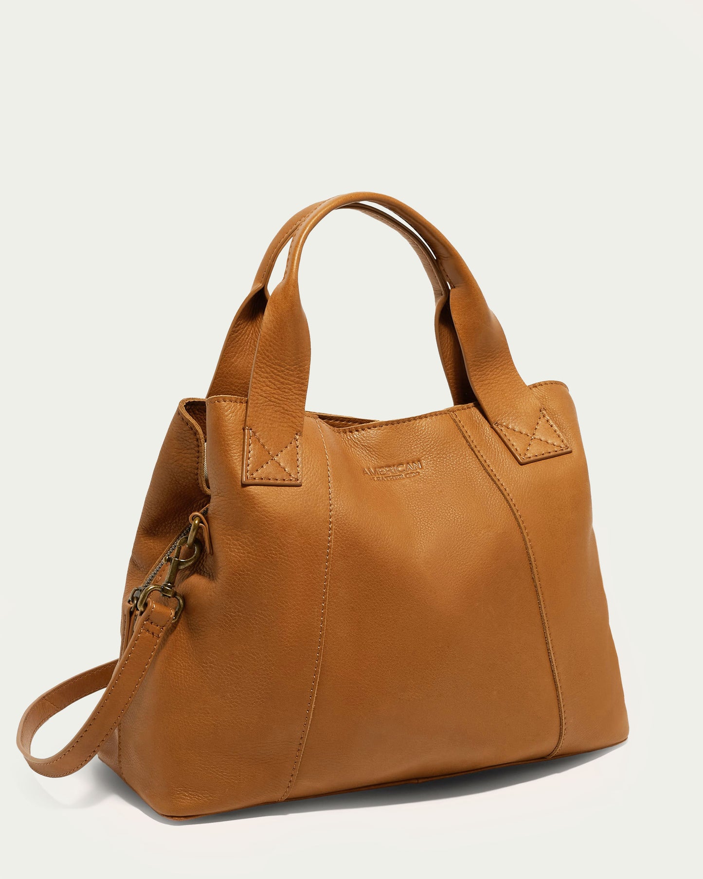 The Ada Triple Entry Satchel by American Leather Co. is a tan leather handbag crafted from authentic American leather, featuring short handles and a detachable crossbody strap. Displayed against a light background, this structured satchel showcases a simple, elegant design with visible stitching accents.