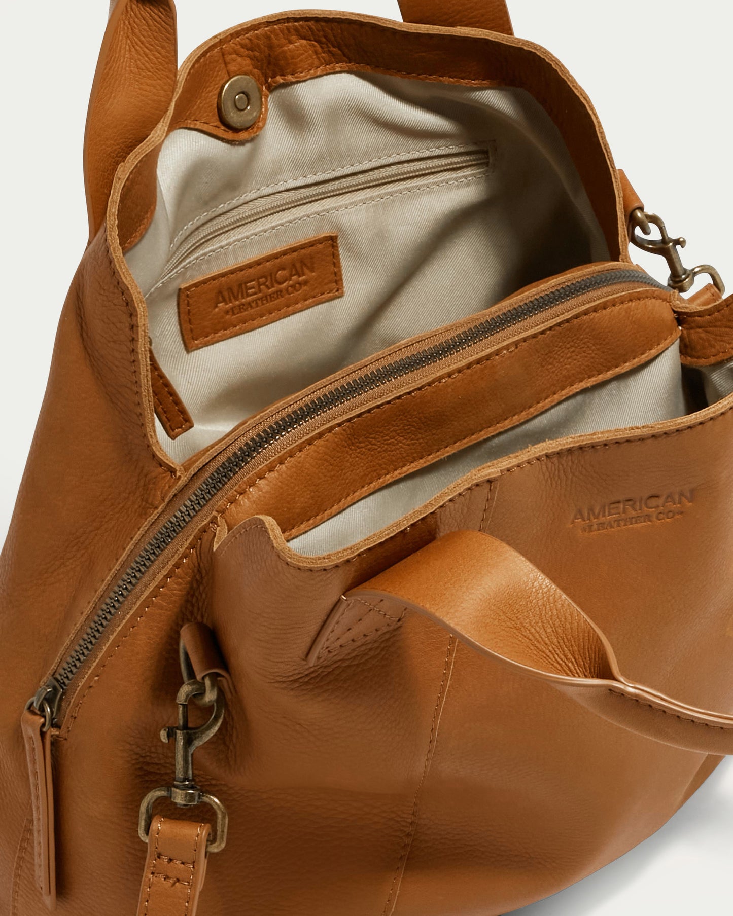 The Ada Triple Entry Satchel by American Leather Co. is an open tan leather handbag featuring visible interior pockets and zippers, along with a tag displaying the brand name. Crafted from genuine American leather, this structured satchel includes a crossbody strap and is elegantly lined with light-colored fabric.