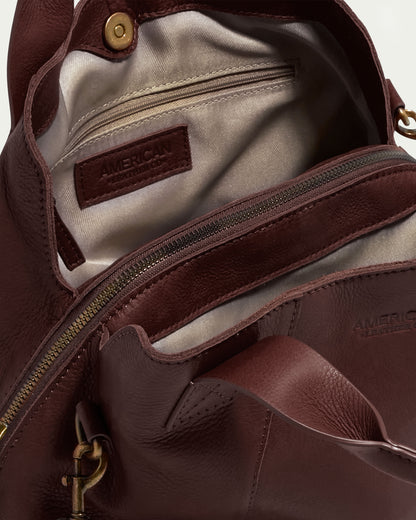 A close-up view of the Ada Triple Entry Satchel from American Leather Co. in brown leather, opened to reveal its interior compartments. Inside, there is a small zip pocket and an open pocket, with the brand name "American Leather Co." embossed on a leather patch inside and on the outer flap. The bag features brass hardware and an adjustable crossbody strap.