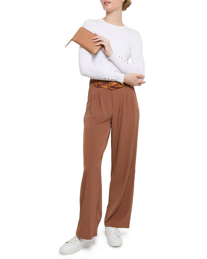 The individual, dressed in a white long-sleeve shirt, brown trousers with a woven belt, and white sneakers, holds an Austin Continental Wallet with RFID by American Leather Co. against a plain white backdrop.