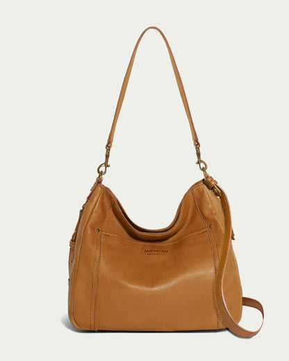 The Austin Convertible Shoulder Crossbody by American Leather Co. is showcased against a light background. This tan leather bag features a slouchy design, an adjustable strap, a front zippered pocket, subtle side pleating, and metal hardware on the shoulder strap. An outside back slip pocket enhances its functionality.