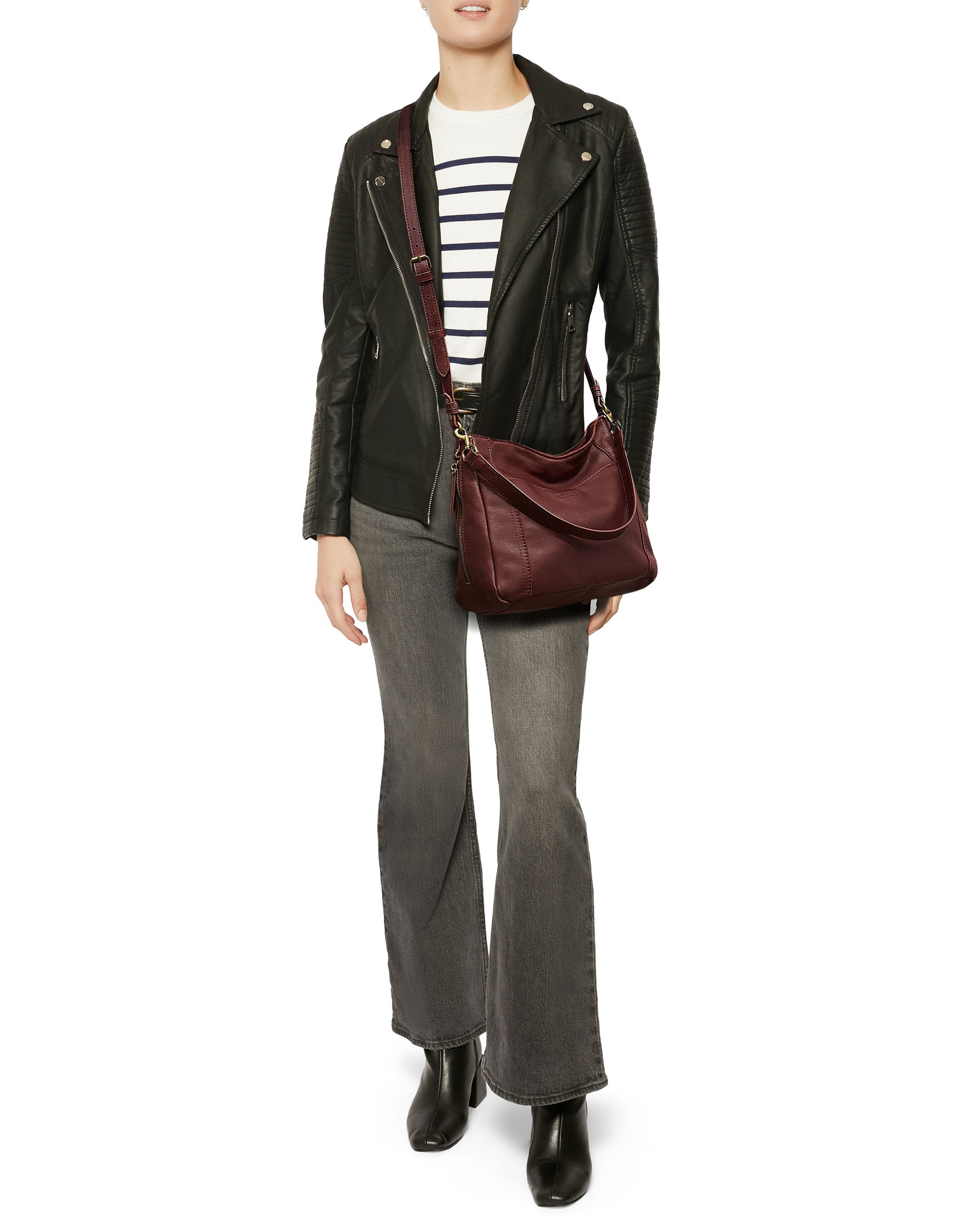 A person is standing, wearing a black leather jacket, a white top with black stripes, gray flared jeans, and black shoes. They are carrying the Austin Convertible Shoulder Crossbody from American Leather Co., which is maroon and has side zipper pockets. Their face is not visible.