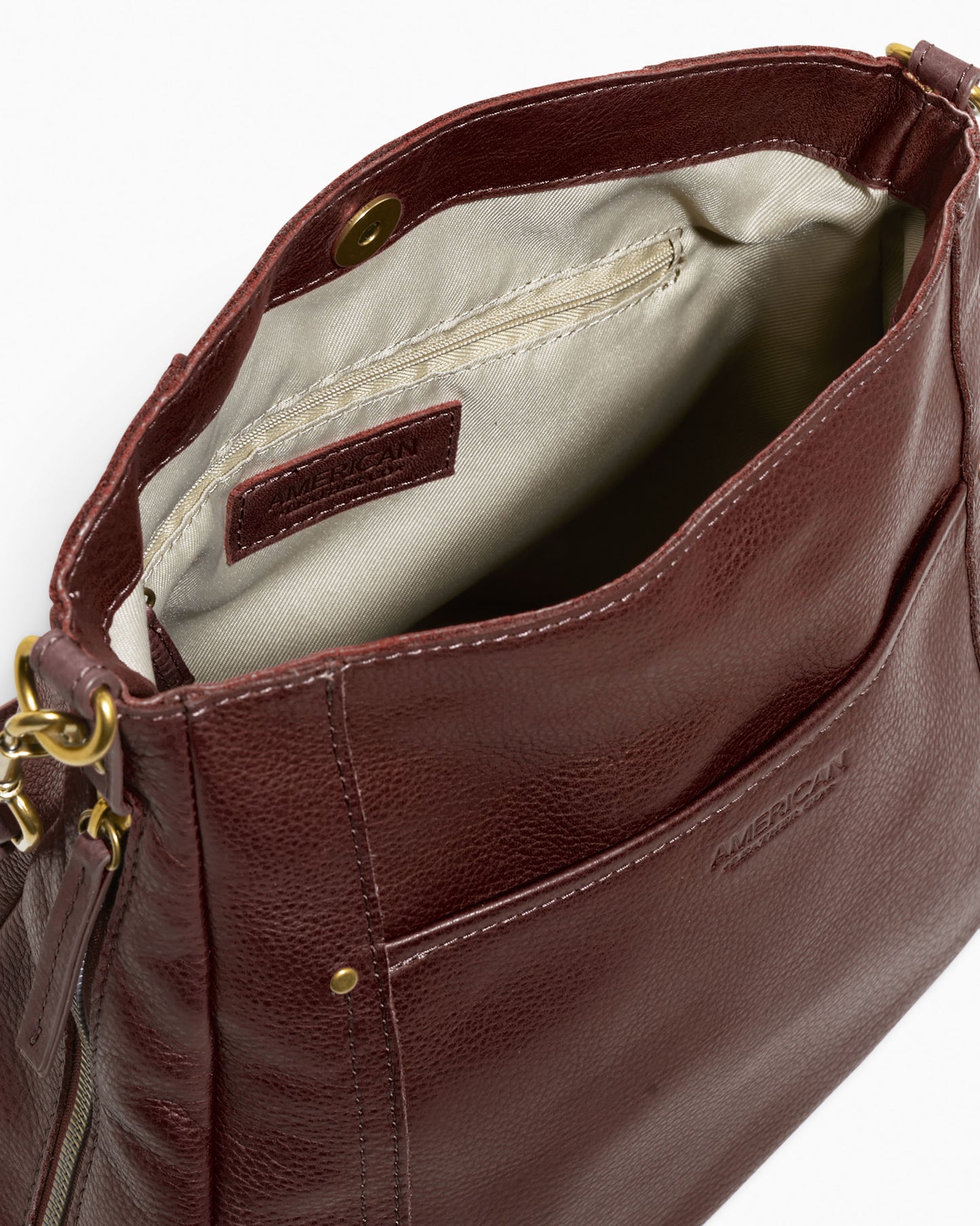 A top-down view of the open Austin Convertible Shoulder Crossbody by American Leather Co. reveals a brown leather exterior and a light beige fabric interior. The bag features a snap button closure, side zipper pockets, and gold-toned hardware. The American Leather Co. logo is visible on the outer pocket and on an inside tag. Additionally, it offers an outside back slip pocket for extra convenience.