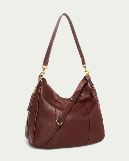 The American Leather Co. Austin Convertible Shoulder Crossbody is a brown leather bag adorned with gold hardware, including buckles and clips. It features a single, adjustable shoulder strap and decorative stitching on its body. Additionally, it has an outside back slip pocket for easy access to essentials. The background is an off-white color.