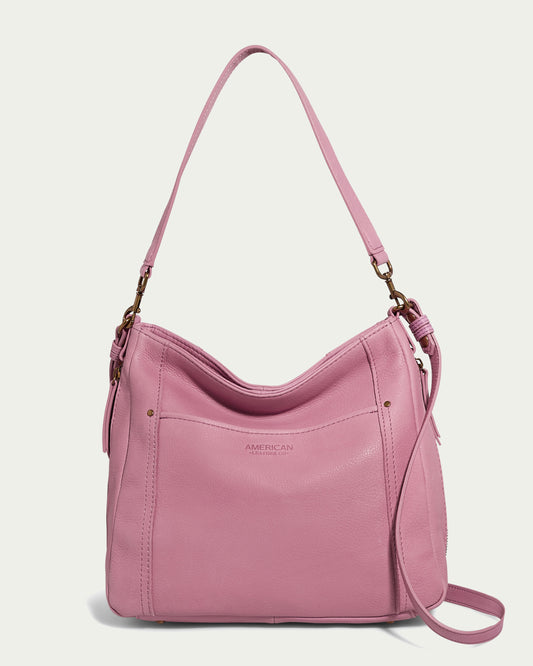 Introducing the Austin Convertible Shoulder Crossbody by American Leather Co., a stylish pink leather bag designed with a shoulder strap, side zipper pockets, and an outside back slip pocket. This elegant piece features subtle stitching and metal hardware, complete with an embossed brand name on the front. Its smooth finish contrasts beautifully against a plain, off-white background.