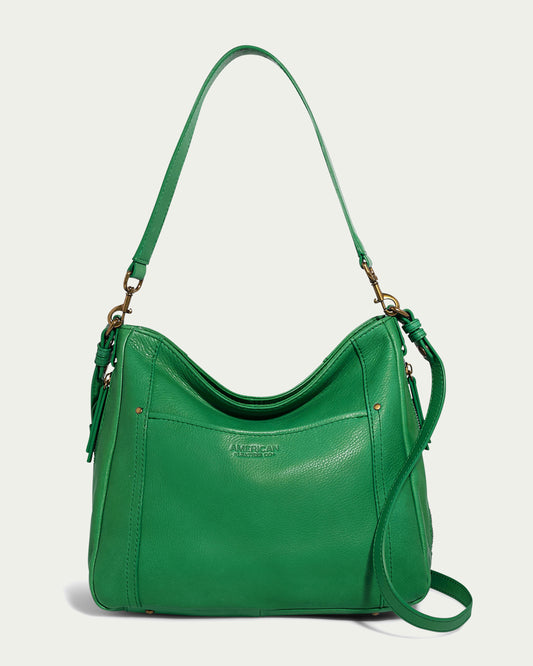 The Austin Convertible Shoulder Crossbody by American Leather Co. is a green leather bag featuring gold hardware and two straps: a short shoulder strap and a longer adjustable crossbody strap. It includes a front pocket, side zipper pockets, and has a slightly slouchy shape. The brand name "American Leather Co." is embossed on the front.