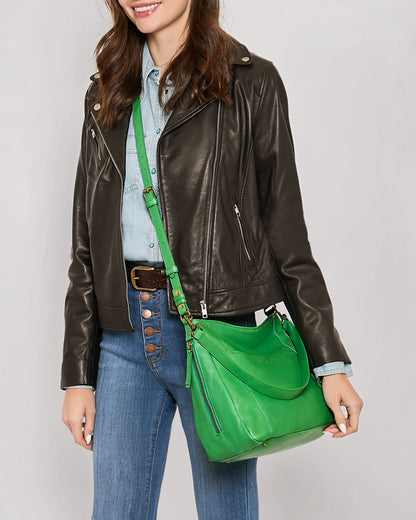 A woman dressed in a black leather jacket and blue jeans is holding the Austin Convertible Shoulder Crossbody from American Leather Co., featuring side zipper pockets. The woman is smiling, and the green leather bag boasts a modern, stylish design with multiple compartments and a smooth finish.