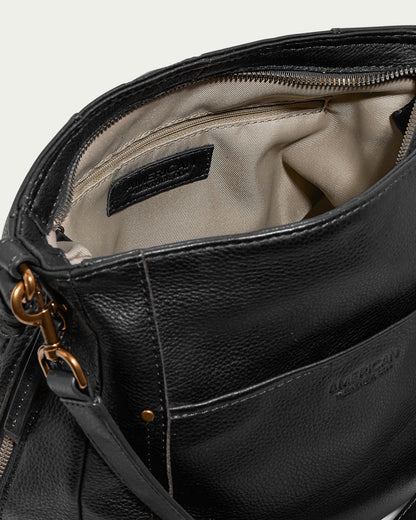 The Austin Convertible Shoulder Crossbody by American Leather Co. is a black leather bag with a brass clasp and a visible interior pocket. The inside is lined with beige fabric, featuring a zippered compartment at the back. Side zipper pockets add extra functionality. The brand "American Leather Co." is embossed on an interior tag and outer pocket.