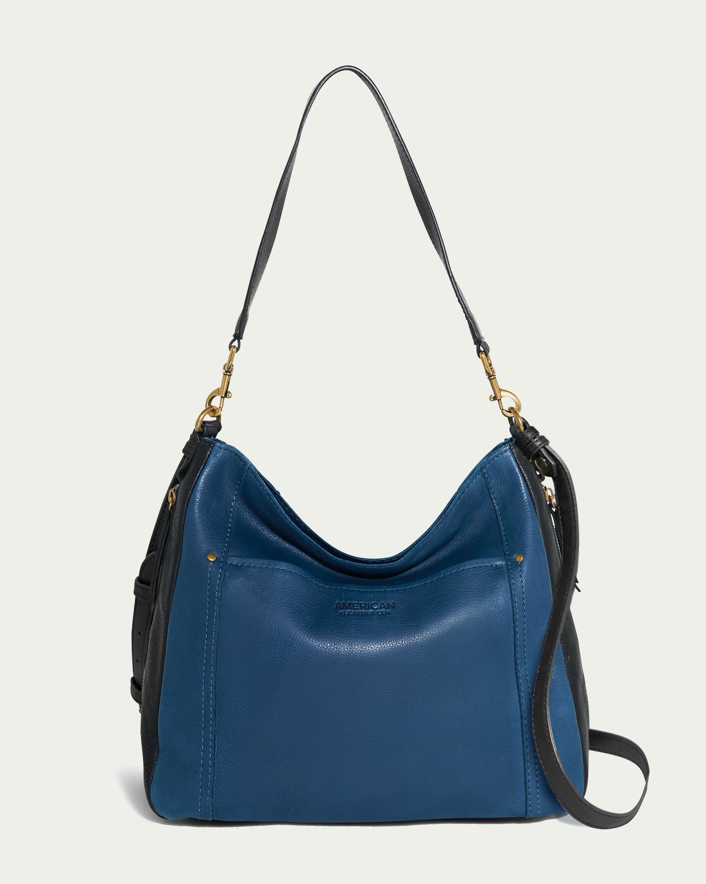 Introducing the American Leather Co. Austin Convertible Shoulder Crossbody, a chic two-toned hobo bag designed with a blue front and black sides. Featuring a convenient shoulder strap and elegant gold-toned hardware, this leather piece includes side zipper pockets and subtle detailing for a contemporary touch.