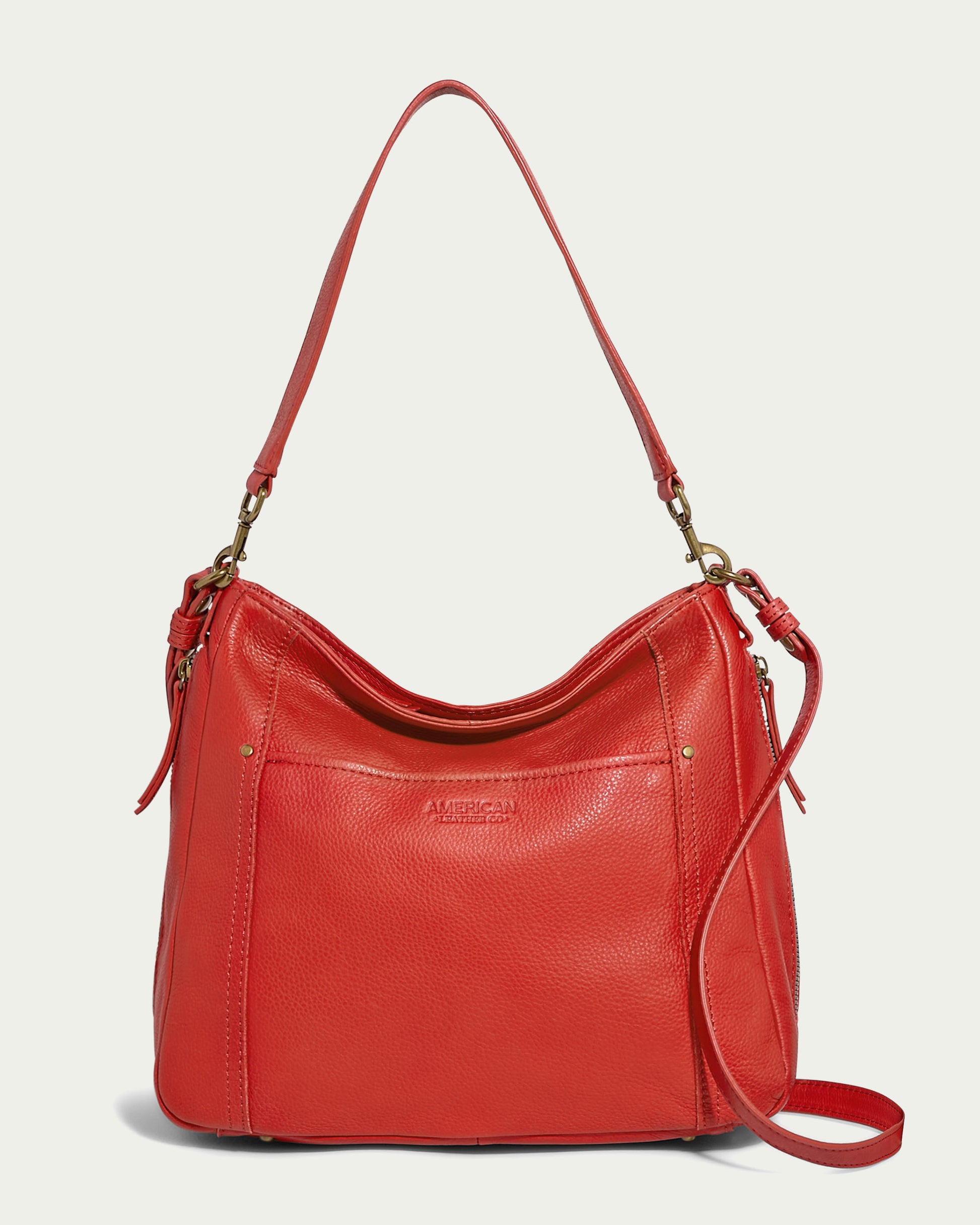 The Austin Convertible Shoulder Crossbody by American Leather Co. is a stylish red leather handbag featuring both a shoulder strap and a smaller crossbody strap. The front showcases a subtle embossed logo along with two small side zipper pockets adorned with gold-tone hardware, making this sleek, rounded bag versatile and elegant.