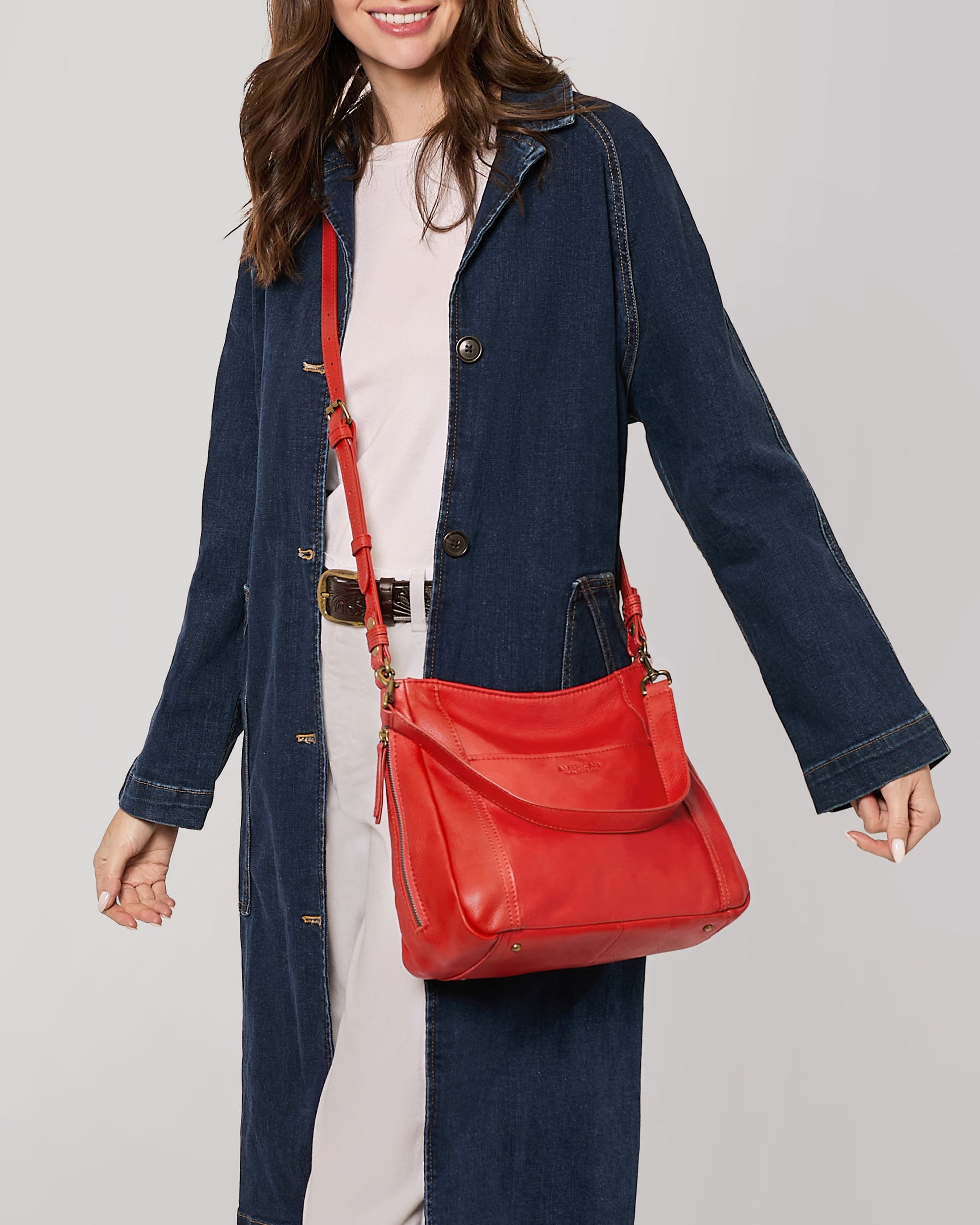 A woman wearing a long denim jacket over a light-colored outfit smiles as she holds the Austin Convertible Shoulder Crossbody from American Leather Co., a red bag featuring an elegant design and convenient side zipper pockets. The handbag also includes a practical shoulder strap. The background is plain and light-colored.