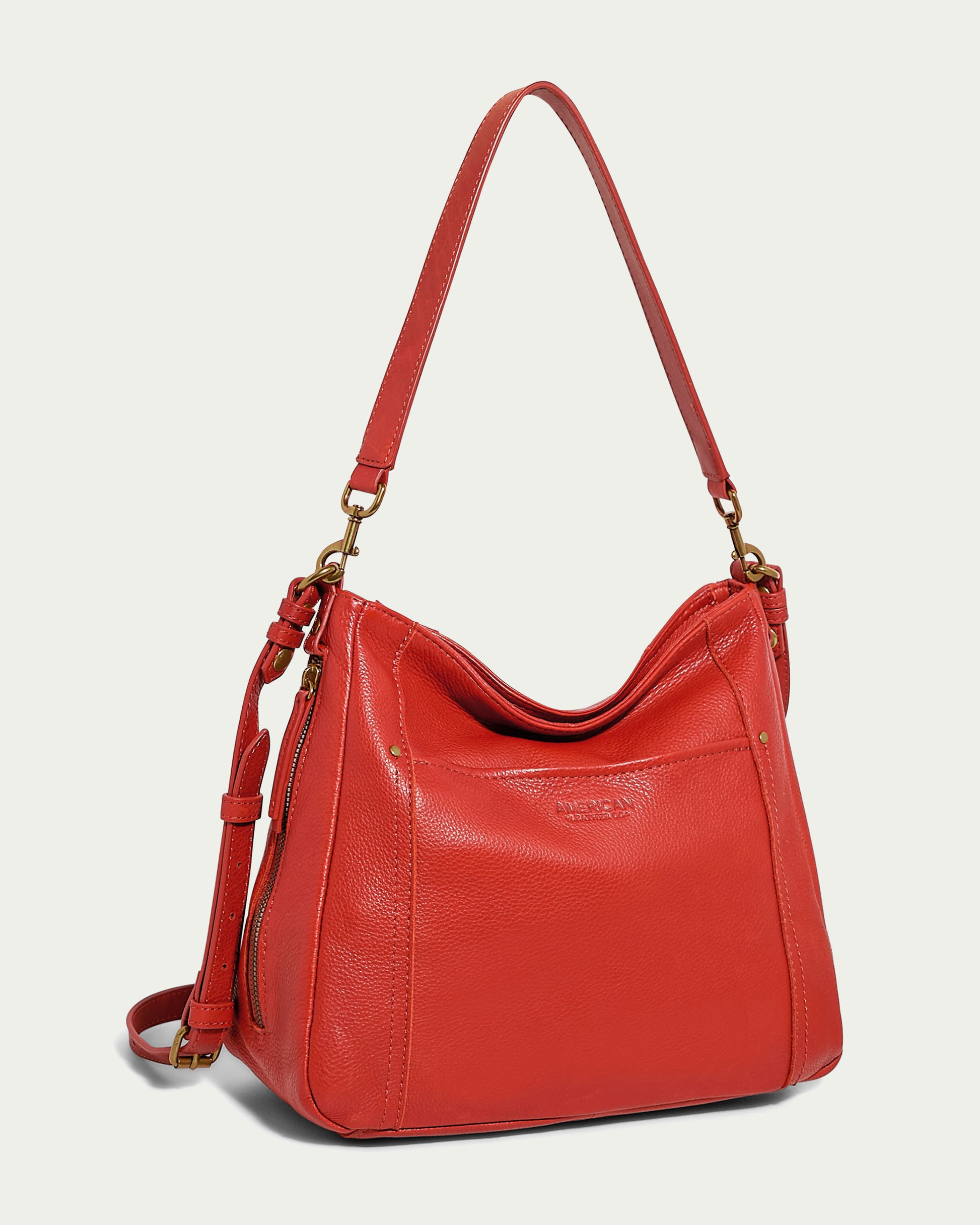 The Austin Convertible Shoulder Crossbody from American Leather Co. is a stylish red leather bag featuring a single adjustable shoulder strap and gold-tone hardware. It boasts a spacious main compartment, side zipper pockets, and the brand logo embossed on the front. Ideal for both casual and semi-formal use.