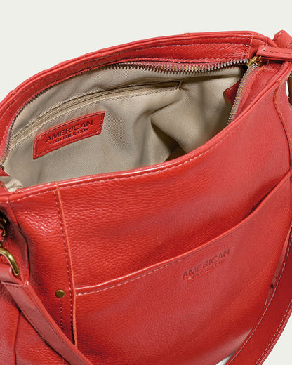 A close-up image of an open Austin Convertible Shoulder Crossbody in red leather reveals a beige interior with a zippered pocket. The brand "American Leather Co." is visible on tags both inside the bag and on the exterior pocket. The bag features gold-tone hardware, side zipper pockets, and a shoulder strap.