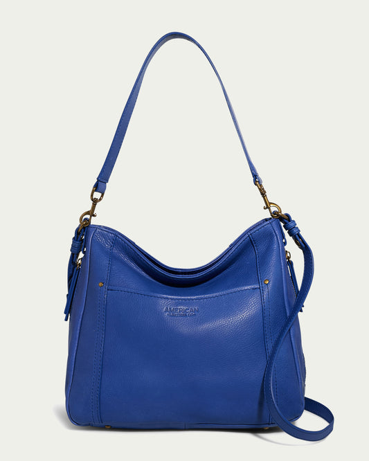 The Austin Convertible Shoulder Crossbody by American Leather Co. is a vibrant blue leather bag with a shoulder strap, brass hardware, and a tassel detail on one side. It features a slightly curved top, front stitching accents, a slip pocket, and the brand name stamped in the center. The background is a light, neutral color.