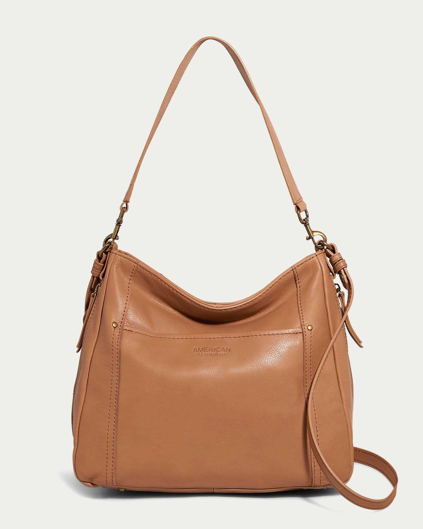 The **Austin Convertible Shoulder Crossbody** from **American Leather Co.** is a tan leather bag featuring both a short shoulder strap and a long, detachable crossbody strap. It showcases minimalistic stitching, a smooth finish, and a small embossed logo at the front center. This elegant bag has zipper pockets and boasts a slightly slouchy shape.