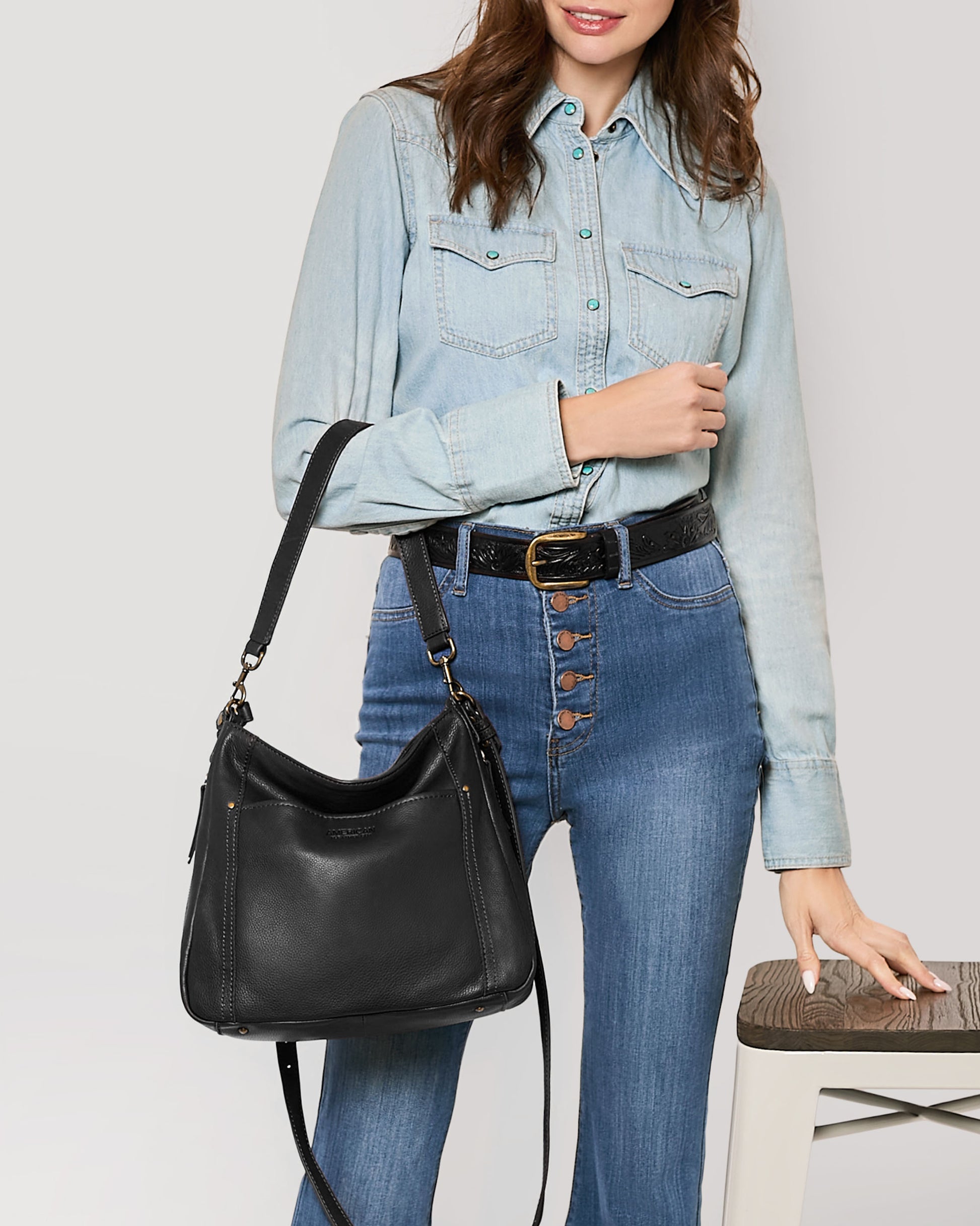 A person wearing a light denim button-up shirt and high-waisted blue jeans stands with an American Leather Co. Austin Convertible Shoulder Crossbody, featuring zipper pockets, over their shoulder. One hand rests on a stool. The outfit is accessorized with a black belt and a brown buckle against a plain, neutral background.