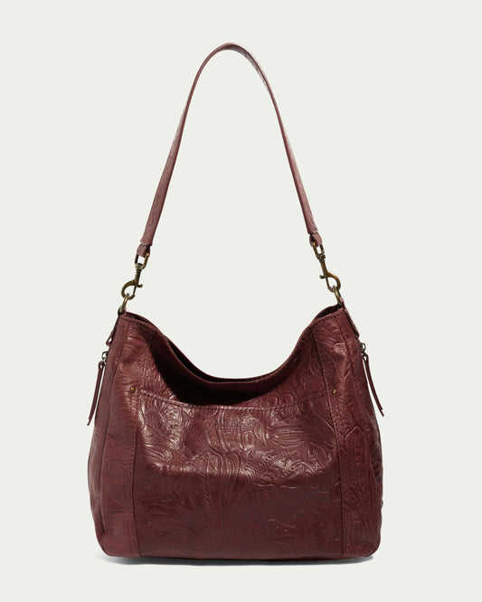 The Austin Shoulder handbag from American Leather Co., made of luxurious maroon leather, features intricate embossed floral designs and a single shoulder strap with metal clasps. This casual accessory, characterized by its slouchy, relaxed shape and small side zippered pockets, is elegantly accented by subtle golden details against a plain, light background.