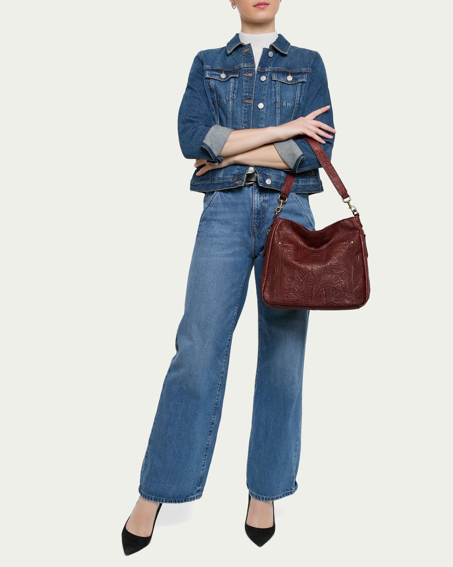 A person is wearing a blue denim jacket over a white shirt, paired with blue denim jeans and black heels. They are holding an Austin Shoulder handbag from American Leather Co. with embossed details. The person's arms are crossed, and their face is not visible.