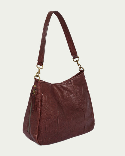 The Austin Shoulder handbag by American Leather Co. is crafted from brown leather and adorned with intricate embossed floral patterns. It features a short strap with gold-colored metal clasps and a convenient side zipper pocket, making it an ideal casual accessory. The bag is beautifully showcased against a plain light background, highlighting its exceptional craftsmanship.