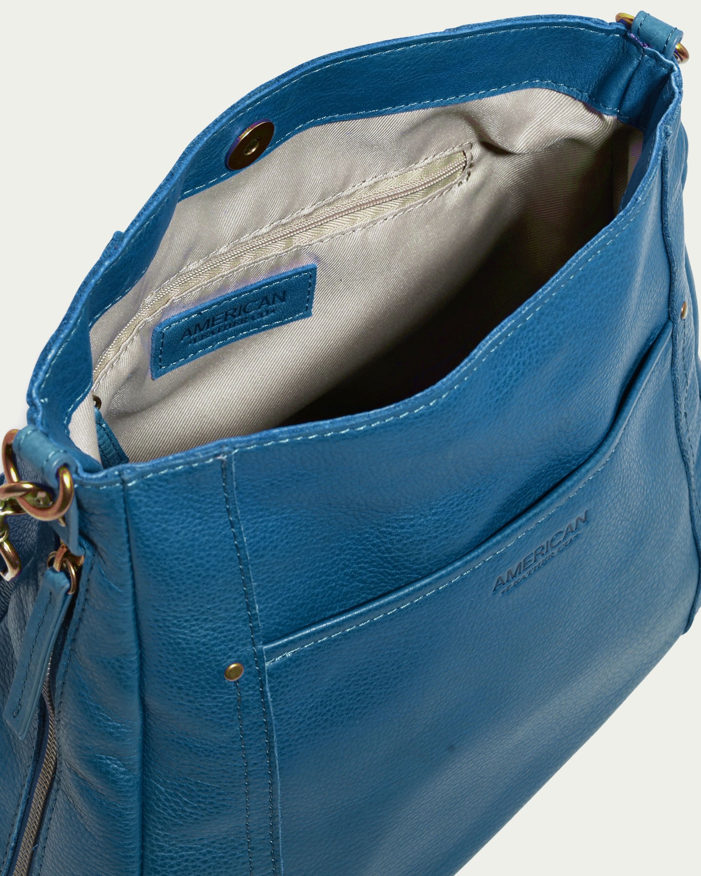 The Austin Shoulder from American Leather Co. is crafted in teal genuine American leather and features an open top that reveals a light beige interior. This casual accessory includes a front pocket, zipper, and metal hardware, with the word "AMERICAN" proudly displayed on the inside label.