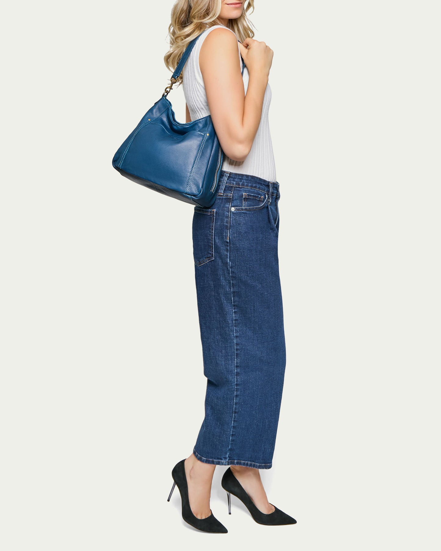 A woman with long blonde hair wearing a white sleeveless top and wide-legged blue jeans stands in profile. She has the Austin Shoulder bag by American Leather Co., made from genuine American leather, draped over her shoulder and wears black high-heeled shoes. The background is plain off-white.