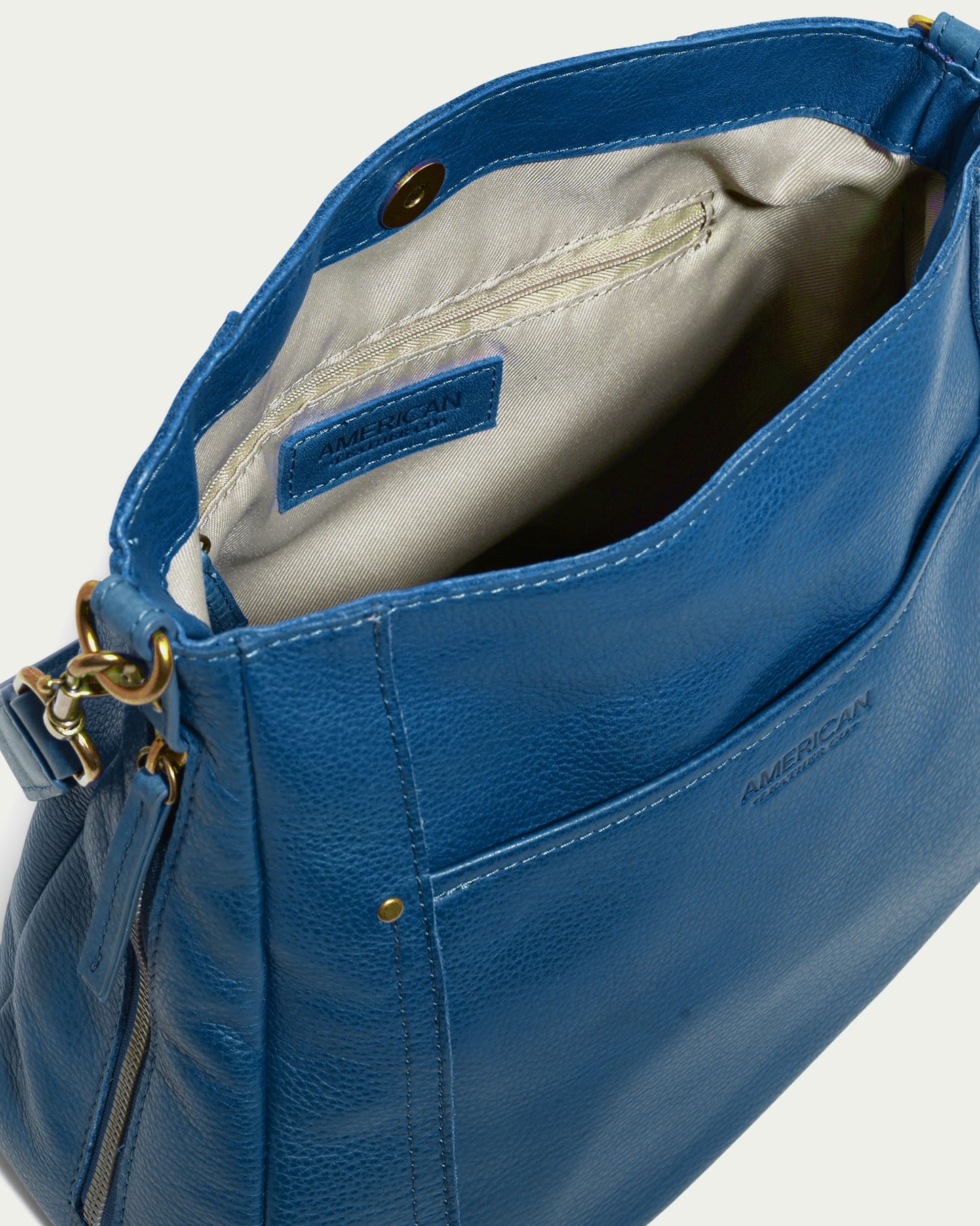 The Austin Shoulder by American Leather Co. showcases a design crafted from genuine American leather. When opened, it reveals a beige interior with a zippered pocket. This bag includes a strap, gold hardware, and an external pocket where the brand's name is prominently embossed on the outer section.