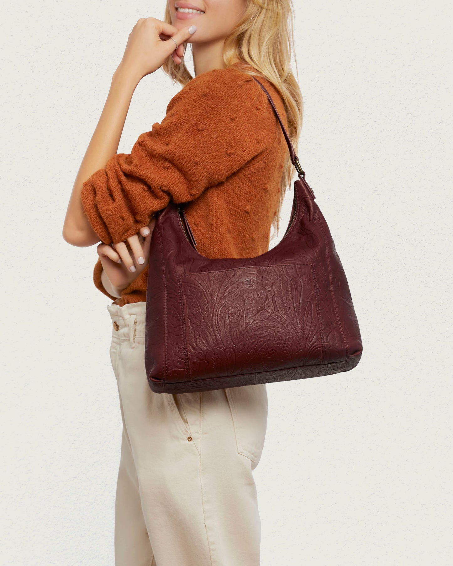 A woman with long blonde hair holds an American Leather Co. Blake Hobo, a large genuine leather burgundy embossed bag. She wears a textured brown sweater and beige pants, with her other hand near her chin against a plain light backdrop.