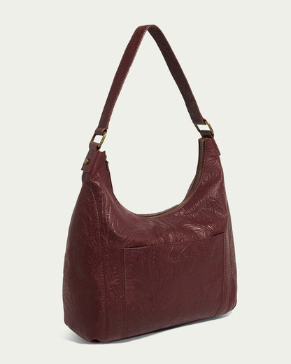 The Blake Hobo by American Leather Co. is a maroon hobo bag made from genuine leather with embossed floral designs. It features a zip closure, front pocket, and single strap with gold-tone hardware against a plain light beige background for added style and functionality.