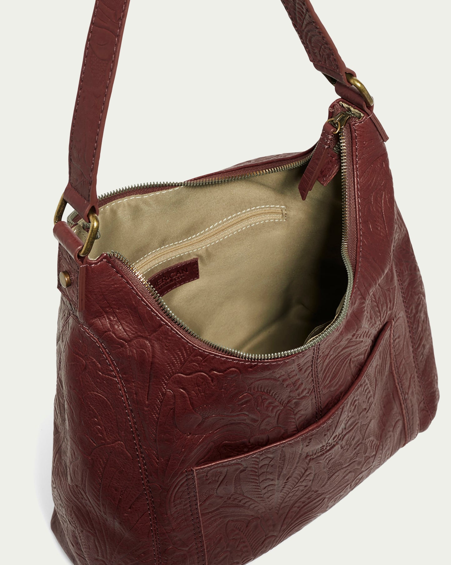 The Blake Hobo by American Leather Co. is a maroon genuine leather bag with floral embossing and a zipper opening. It includes a beige fabric lining, interior pocket, strap, antique brass hardware, and exterior open pocket for convenience.