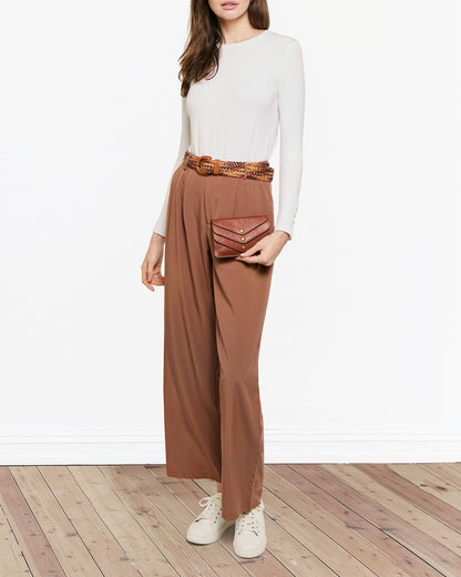 A woman in a white long-sleeve shirt and brown wide-leg pants stands on a wooden floor, wearing white sneakers and a woven belt, while holding an American Leather Co.'s Budd Multi-Pocket Wallet With RFID, which doubles as an envelope-style clutch.