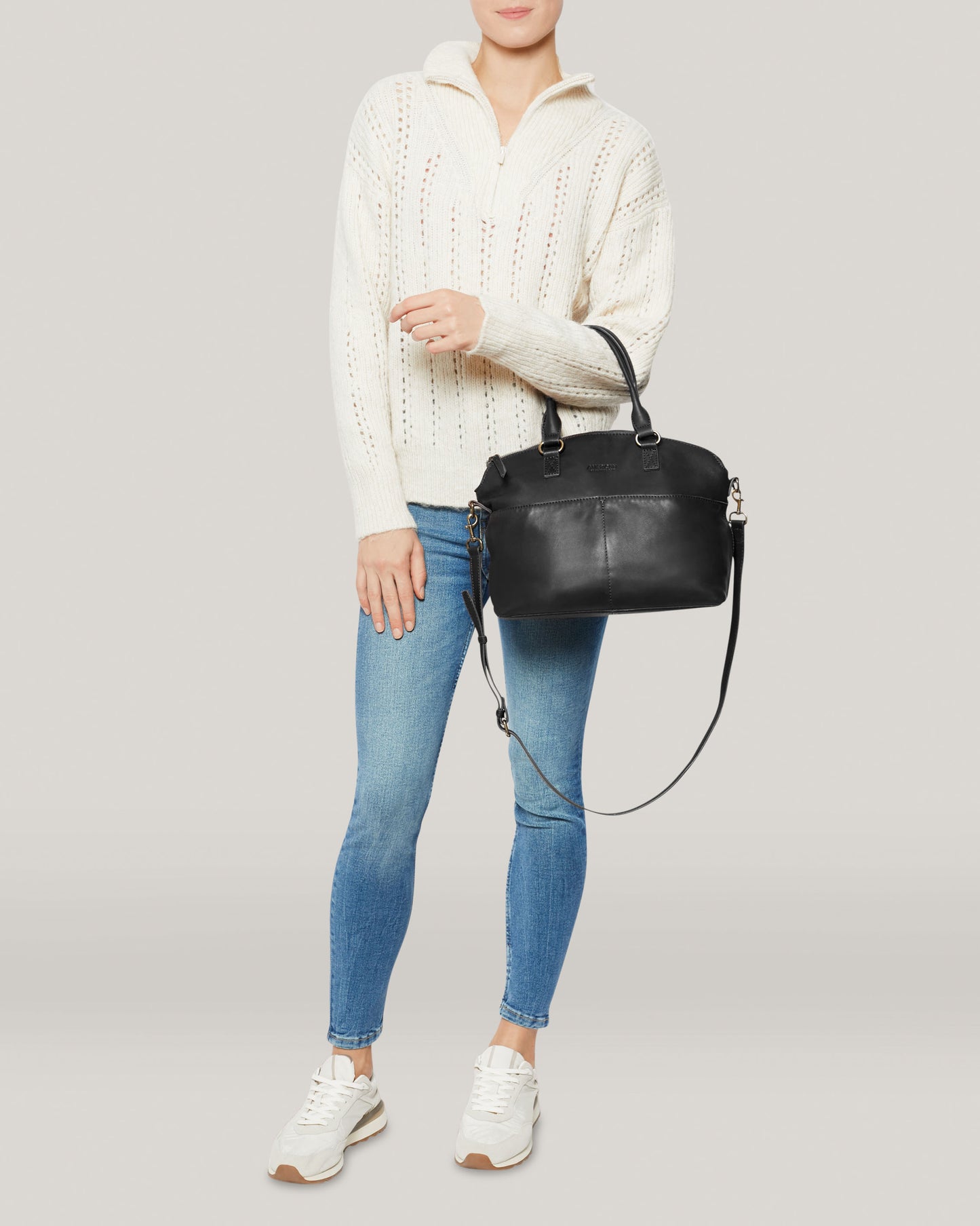 A woman wearing a cream sweater and blue jeans holds the Carrie Dome Satchel by American Leather Co., which comes with an adjustable crossbody strap. She completes her casual look with white sneakers while standing against a light background, with the genuine leather bag adding a touch of elegance.