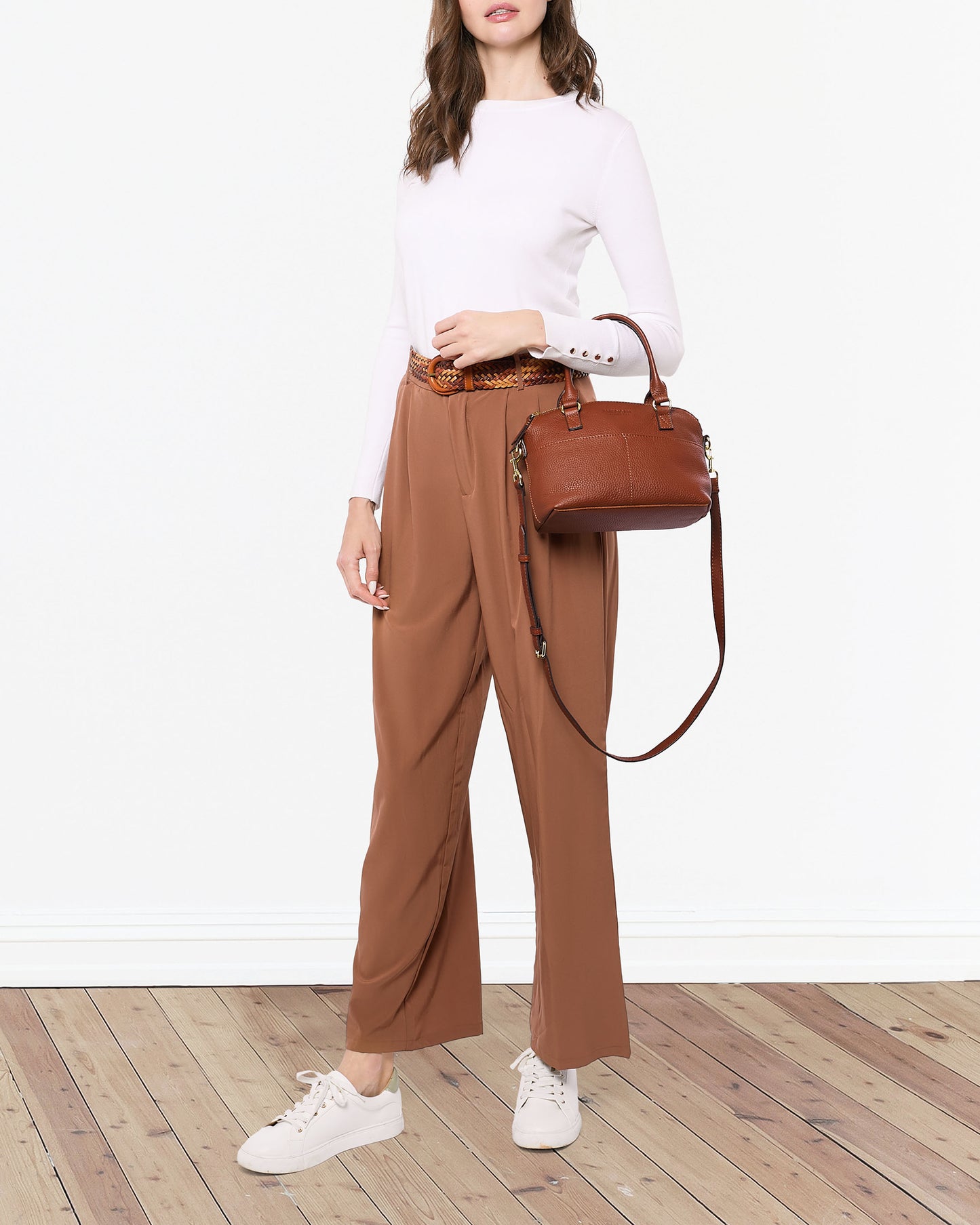 A woman stands on a wooden floor, wearing a white long-sleeve top, tan high-waisted pants, and white sneakers. She holds the Carrie Mini Dome Crossbody bag from American Leather Co., made from genuine leather with a shoulder strap. Her hair is down, and she is posing with one hand on her hip against a plain white wall.