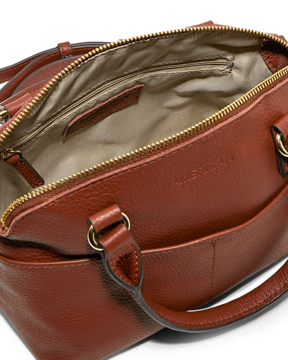 A brown leather Carrie Mini Dome Crossbody handbag from American Leather Co. reveals a beige fabric-lined interior featuring both a zippered pocket and an open pocket. The exterior showcases embossed "AMERICAN LEATHER CO." text and gold-toned hardware, including two ring attachments for the handles.