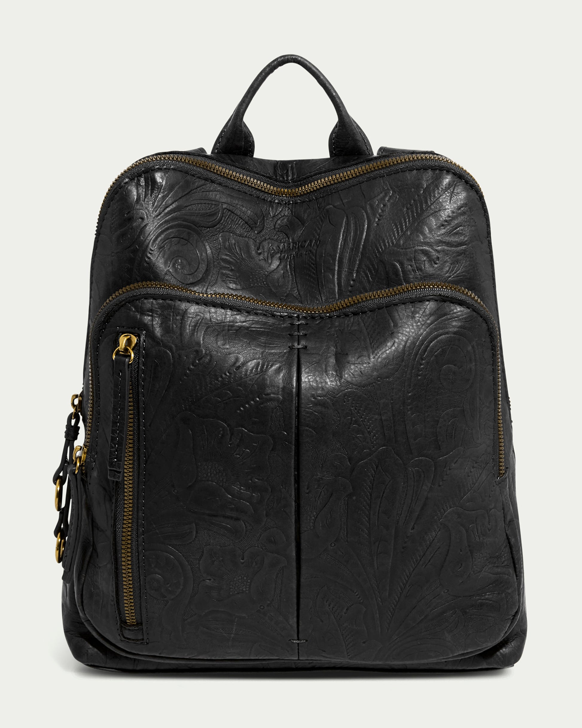 The Cleveland Backpack by American Leather Co. is a black tooled leather backpack featuring embossed floral patterns, two front zippered pockets with gold-tone zippers, a top handle, and adjustable straps. It maintains a structured shape with subtle ornamental detailing.