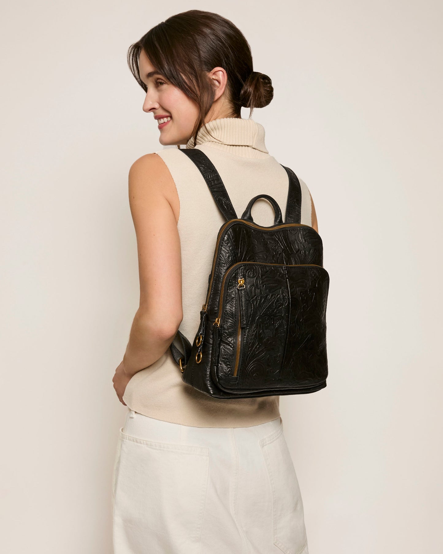 A dark-haired woman wears a sleeveless beige turtleneck and white pants, carrying the Cleveland Backpack by American Leather Co. Made from genuine American leather with embossed designs, its adjustable straps enhance style and comfort. She smiles and looks right against a plain backdrop.