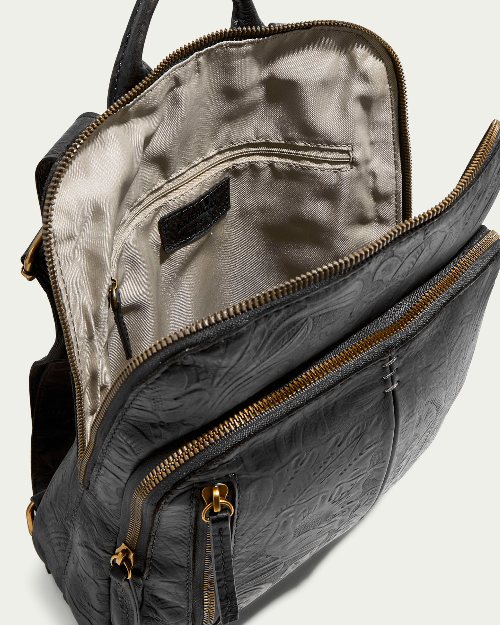 The Cleveland Backpack by American Leather Co. is crafted from genuine black leather with an open top revealing a beige interior. It features embossed patterns, multiple compartments such as a front zip pocket, gold-toned zippers, and adjustable straps against a light background.