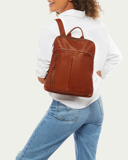 A person with curly hair, wearing a white shirt and blue jeans, is seen from the back, smiling and carrying an American Leather Co. Cleveland Backpack made of genuine American leather over their left shoulder. The Cleveland Backpack has multiple compartments, a sleek design, and adjustable back straps.