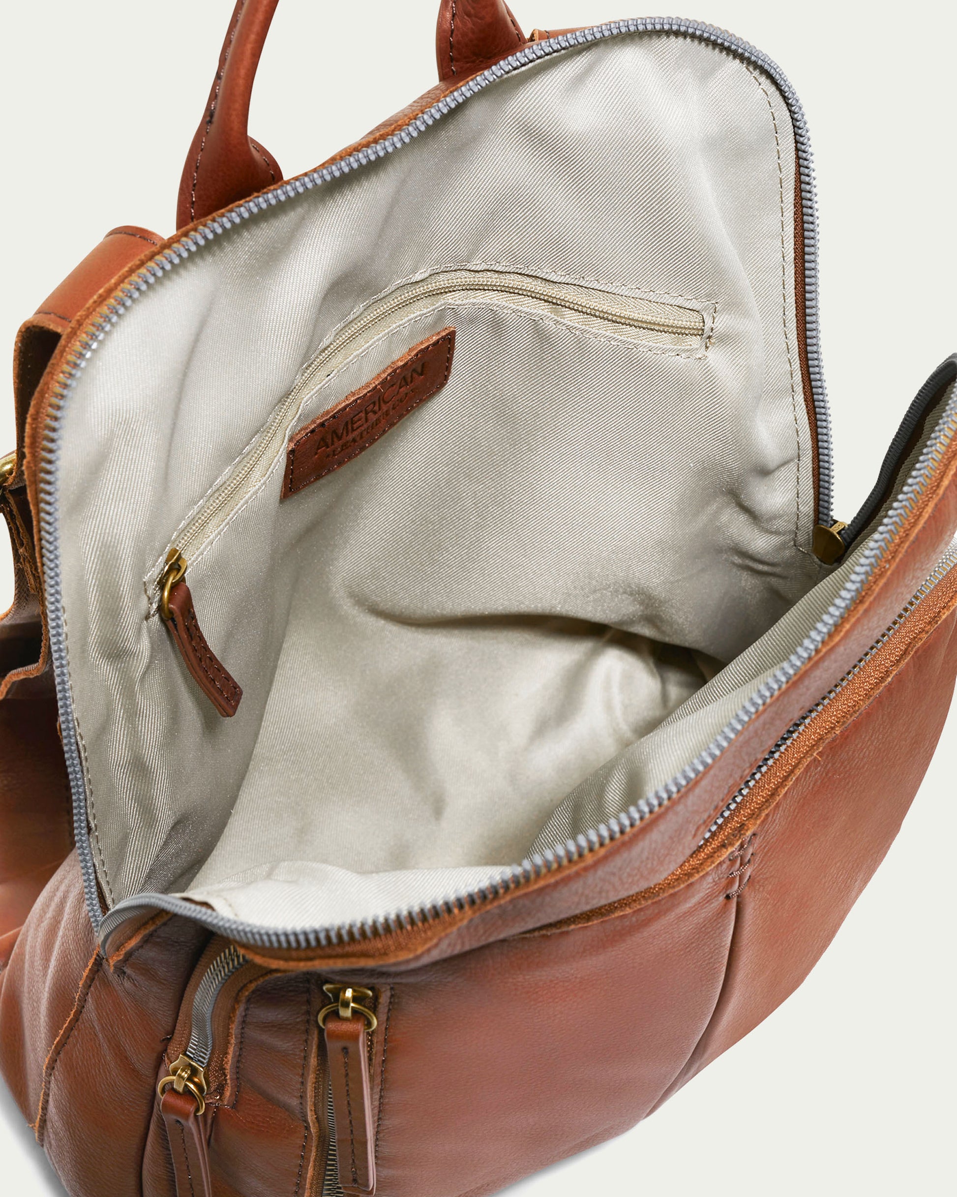 The Cleveland Backpack by American Leather Co. is a brown leather backpack purse featuring adjustable back straps and a light-colored fabric inner lining. The backpack is shown open, revealing multiple interior compartments with the zipper partially open, showcasing its spacious inside and zippered pocket for organization. It's crafted from genuine American leather.
