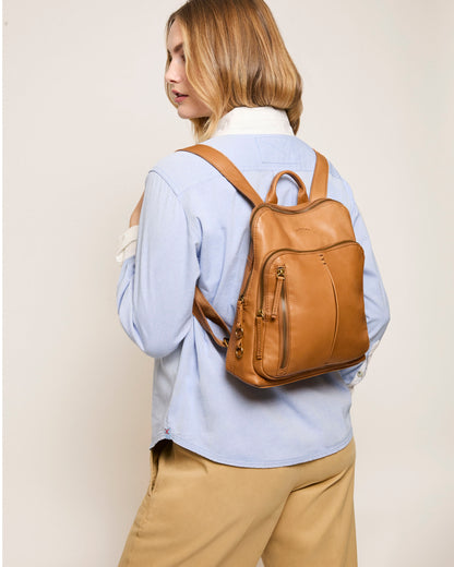 In a light blue shirt and beige pants, a person with long hair stands with their back turned, showcasing the Cleveland Backpack by American Leather Co. It's crafted from genuine American leather and features adjustable straps, set against a plain light-colored background that highlights its simplicity.